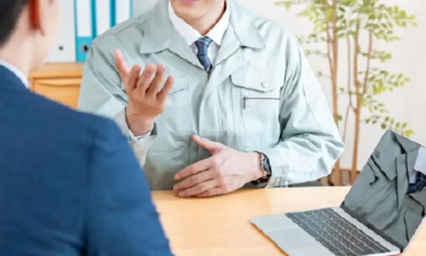作業服を着た社員がスーツ姿の人と面談している様子。ノートパソコンも机の上にある。